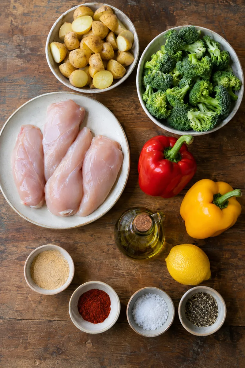 Ingredients laid out: chicken breasts, baby potatoes, broccoli, bell peppers, olive oil, spices and lemon