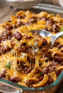Frito chili pie bake in a glass dish topped with melted cheddar and corn chips, spoon lifting a cheesy scoop